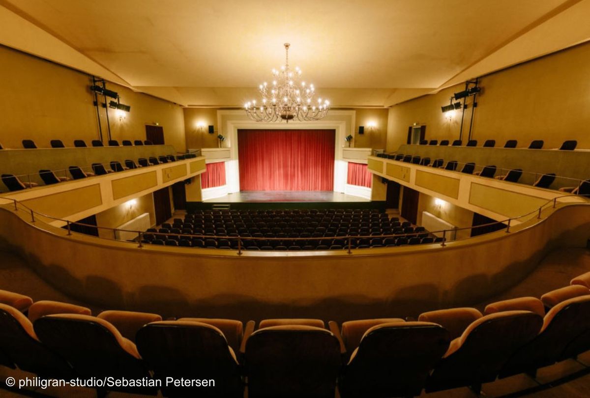 Saal des Brunnentheater mit Blick auf die Bühne mit roten Vorhang und den großen Kronleuchter